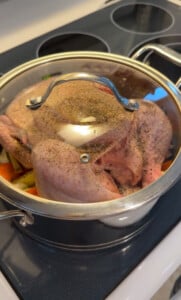 A whole raw chicken seasoned with pepper sits in a pot over chopped vegetables, covered with a glass lid, on a stovetop—perfect for making No Water Chicken Soup, a waterless chicken soup recipe that captures pure flavor.