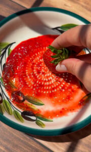 A hand holding a fresh strawberry is dipping it into a decorative plate with a spiral pattern of red strawberry sauce beside soft rice paper mochi on a wooden surface.
