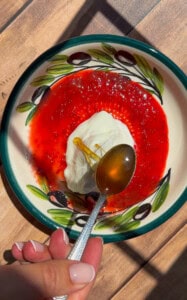 A hand holds a spoon with honey over a bowl containing white yogurt, rice paper mochi, and bright red fruit sauce, placed on a wooden surface with sunlight and shadow patterns.