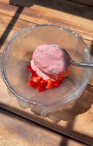 A spoonful of creamy pink yogurt with strawberry pieces in a glass bowl, accompanied by delicate rice paper mochi, sits on a wooden surface in bright sunlight.