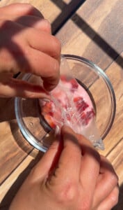 Two hands peel off a thin, clear, plastic-like sheet—resembling rice paper mochi—from a glass bowl containing small pieces of red fruit, possibly strawberries, on a wooden surface in natural sunlight.