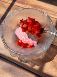A glass bowl with pink yogurt topped with chopped fresh strawberries sits on a wooden surface, alongside delicate rice paper mochi. A spoon holds more chopped strawberries above the bowl as sunlight casts gentle shadows on the table.
