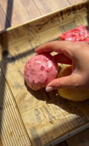 A hand holding a round, pink, berry-studded ice cream scoop on a tray lined with printed rice paper mochi parchment, with sunlight casting soft shadows.