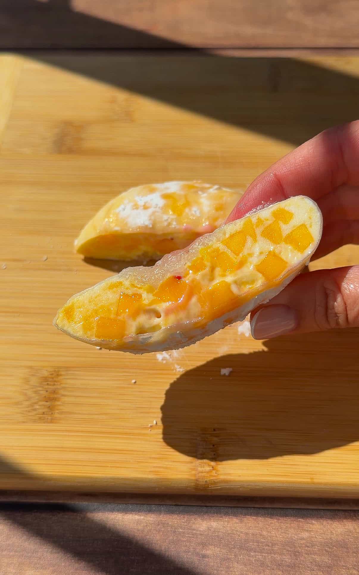 A hand holds half of a rice paper mochi dessert filled with creamy white and orange mango pieces on a wooden cutting board, with sunlight casting soft shadows.