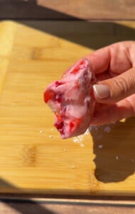 A hand with light-colored nails holds a bitten piece of strawberry rice paper mochi ice cream over a wooden cutting board in sunlight.