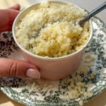 A hand holds a small cup filled with cooked pasta topped with grated cheese and black pepper, placed on a decorative green and white plate. A spoon is in the cup, and some cheese is scattered on the plate.