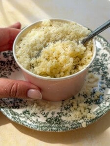 A hand holds a small cup filled with cooked pasta topped with grated cheese and black pepper, placed on a decorative green and white plate. A spoon is in the cup, and some cheese is scattered on the plate.