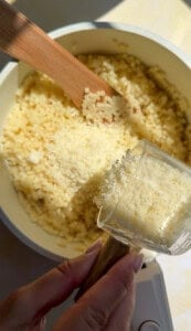 A hand holds a measuring cup filled with grated cheese over a pot of risotto, while a wooden spoon stirs the creamy rice mixture in the sunlight.