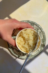 A hand holds a small cup filled with star-shaped pasta in a creamy sauce, placed on a decorative green and white saucer. A spoon rests inside the cup, with sunlight casting shadows on the surface.