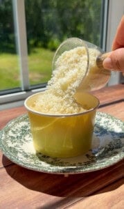 A hand pours grated cheese from a small cup into a yellow paper bowl, which sits on a decorative plate by a sunlit window with a green outdoor view.