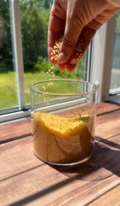 A hand sprinkles small yellow beads into a clear glass container, which is already partially filled, sitting on a sunlit wooden surface by a window with a garden view.