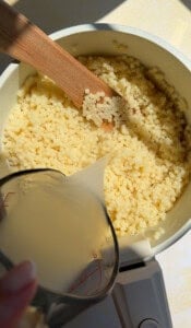 A close-up of a pot with small pasta being stirred by a wooden spoon, while a person pours liquid from a measuring cup into the pot. Bright sunlight illuminates the scene.