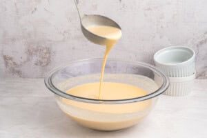 A ladle pours smooth, pale yellow custard batter back into a glass bowl, with two stacked white ramekins in the background on a light surface.