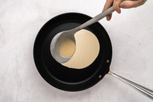 A hand pours pancake batter from a ladle into a black nonstick frying pan on a light-colored surface.