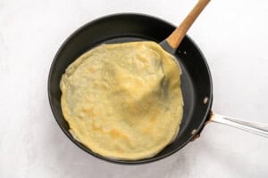 A thin crepe cooking in a nonstick skillet is being lifted at the edge with a spatula. The background is a light, neutral surface.