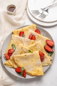 A plate of folded crepes topped with sliced strawberries, mint leaves, and powdered sugar. A small jug of syrup, stacked plates, and forks are nearby on a light-colored tablecloth.
