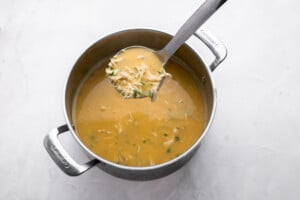 A stainless steel pot filled with creamy yellow soup and shredded chicken, with a ladle lifting some soup out, shown on a white background.