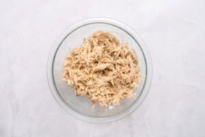 A clear glass bowl filled with shredded cooked chicken sits on a light marble surface.