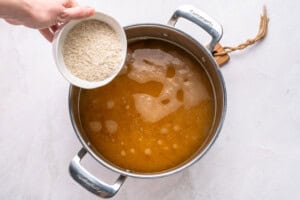 A hand holds a white bowl of uncooked rice over a pot filled with simmering broth on a white surface, preparing to add the rice to the pot.