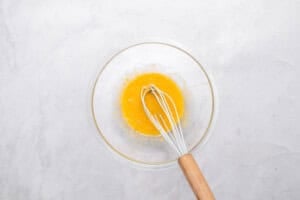 A clear glass bowl with a wooden-handled whisk mixing beaten eggs on a light gray marble surface.