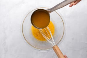 A hand pours orange liquid from a ladle into a glass bowl with a whisk, likely preparing a mixture, on a light countertop.