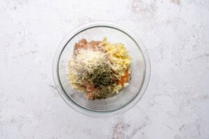 A glass bowl containing ground meat, grated cheese, chopped herbs, shredded cheese, and seasonings on a light-colored surface.