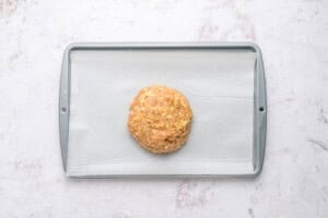 A ball of uncooked dough sits on a parchment-lined baking sheet against a light, textured background.
