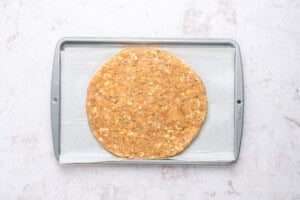 A raw, round mixture of seasoned ingredients is flattened on parchment paper atop a metal baking sheet, ready to be baked, against a light-colored textured background.