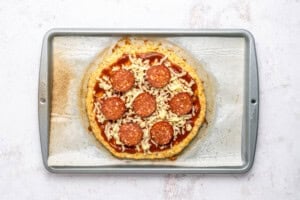 A homemade pepperoni pizza with shredded cheese and tomato sauce sits on parchment paper on a baking tray, ready to be baked.