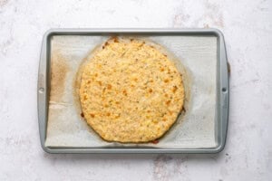 A baked round cauliflower pizza crust sits on parchment paper on a metal baking sheet, viewed from above on a light textured surface.