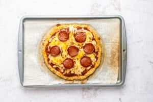 A baked pepperoni pizza with melted cheese sits on parchment paper on a metal baking sheet, viewed from above on a light-colored surface.