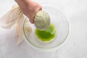 A hand squeezes a cloth bundle over a glass bowl, extracting green liquid from the bundle onto a white surface.