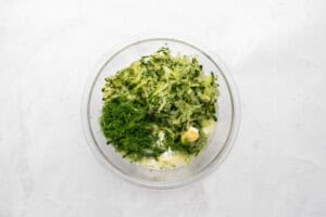 A glass bowl containing grated cucumber, chopped fresh dill, minced garlic, and a dollop of yogurt or sour cream on a white surface.