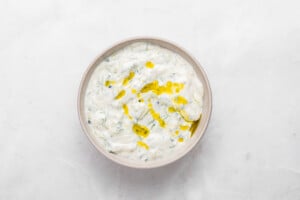 A bowl of creamy tzatziki sauce garnished with a drizzle of olive oil, set on a white marble surface.