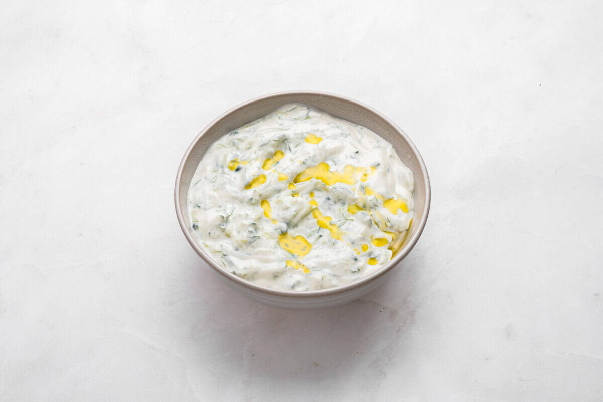 A bowl of creamy yogurt dip mixed with herbs, topped with a drizzle of olive oil, set on a light, textured surface.