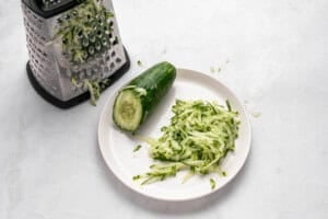 A plate with shredded cucumber next to a half cucumber, with a metal box grater covered in cucumber shreds on a white surface.