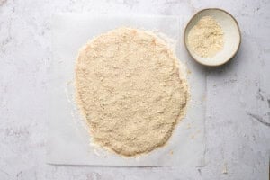 A sheet of parchment paper with rolled-out dough covered in crumbled mixture sits beside a bowl filled with more crumbs on a light-colored surface.