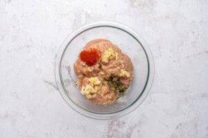 A glass bowl containing ground meat mixed with red seasoning, minced garlic, and dried herbs, placed on a light, textured surface.