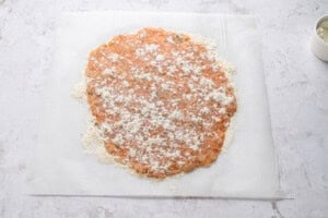 Raw ground meat mixture rolled out flat on parchment paper, sprinkled with flour. A small white ramekin sits in the upper right corner on a light, textured surface.