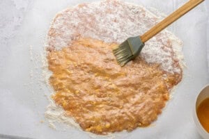 A pastry dough rolled out on parchment paper is being brushed with an egg wash using a silicone pastry brush. Flour is visible around the edges of the dough.