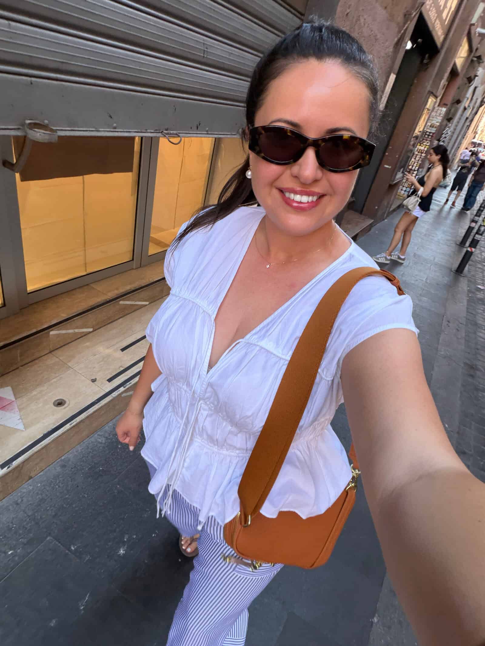 A woman wearing sunglasses, a white blouse, and striped pants takes a selfie while walking down a city street. She is smiling and carrying a brown shoulder bag. Shops and people are visible in the background.