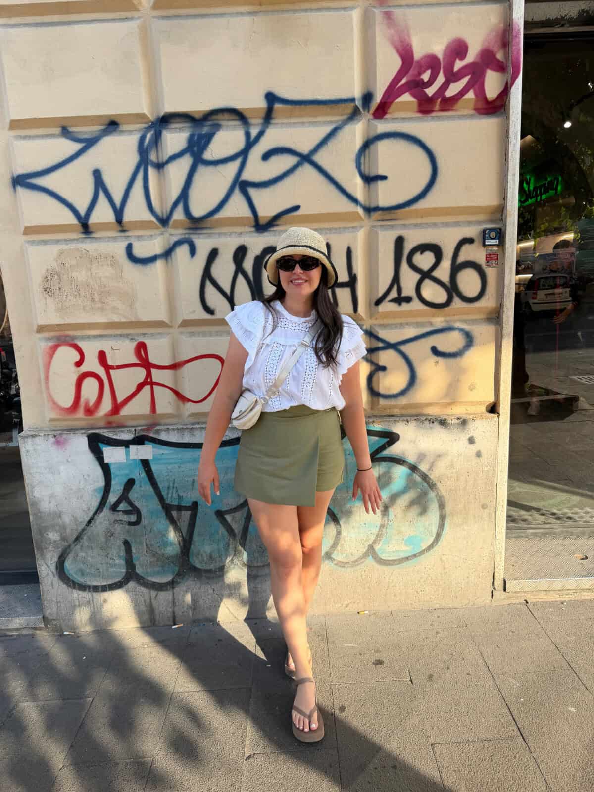 A woman wearing a white blouse, green shorts, sandals, a hat, and sunglasses stands smiling in front of a graffiti-covered wall on a sunny day.