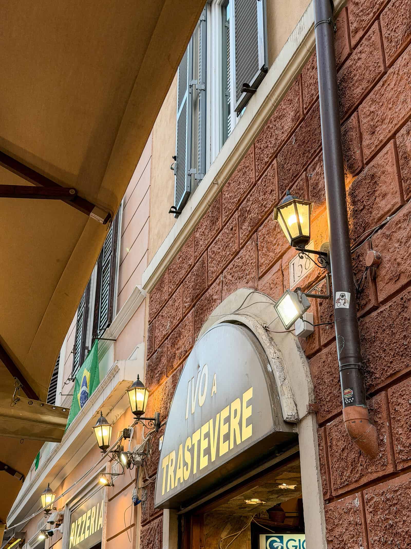 A street view of a building in Rome with a sign reading "IVO A TRASTEVERE" above an arched entrance, illuminated by wall lanterns, and an adjacent "PIZZERIA" sign visible to the left.