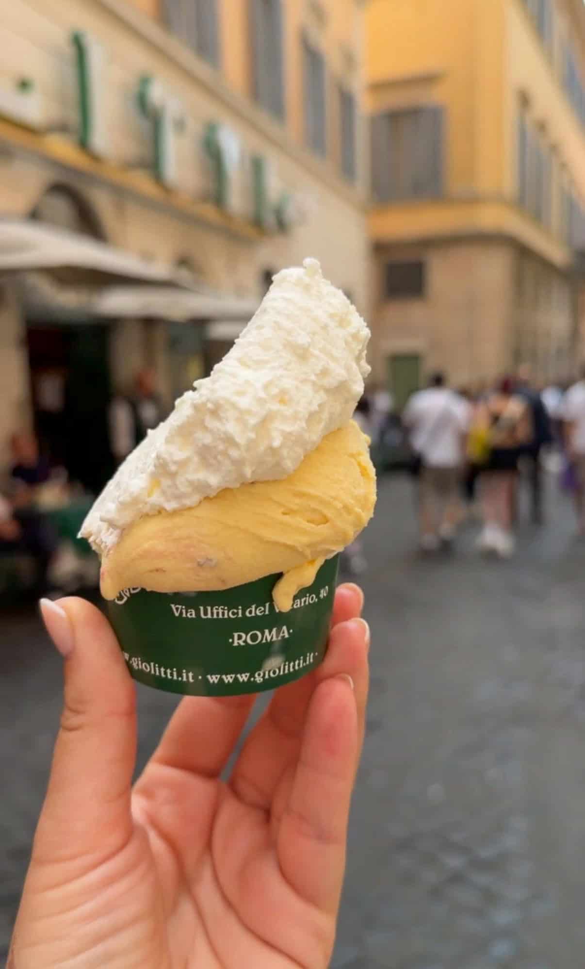 A hand holds a cup of gelato with a large swirl of whipped cream on top; blurred pedestrians and yellow buildings are visible in the background, suggesting a lively street scene in Rome.