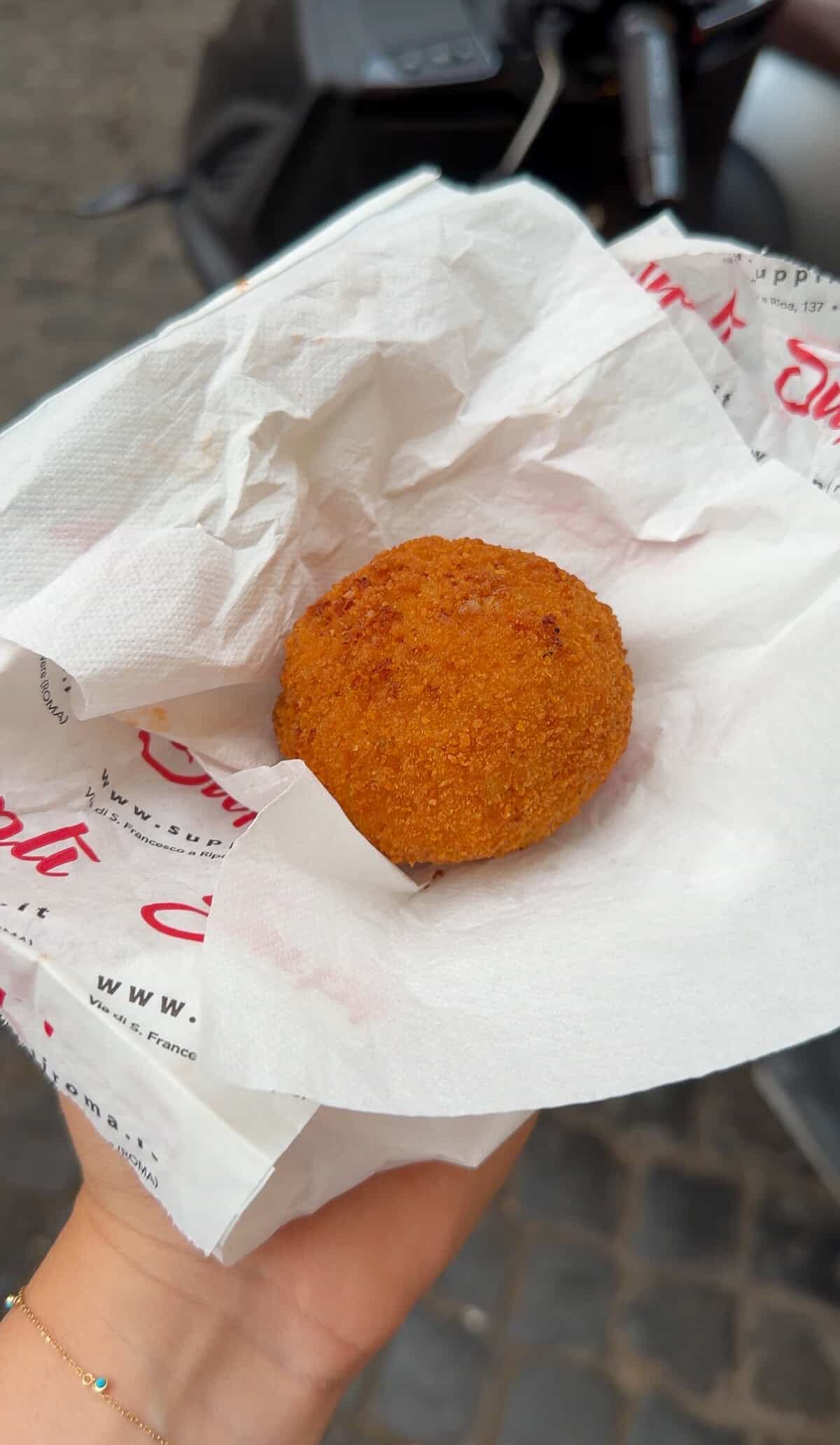 A hand holds a single golden-brown, breaded and fried ball, resting on a white napkin with printed text. The background is blurred, showing cobblestones and part of a dark object.
