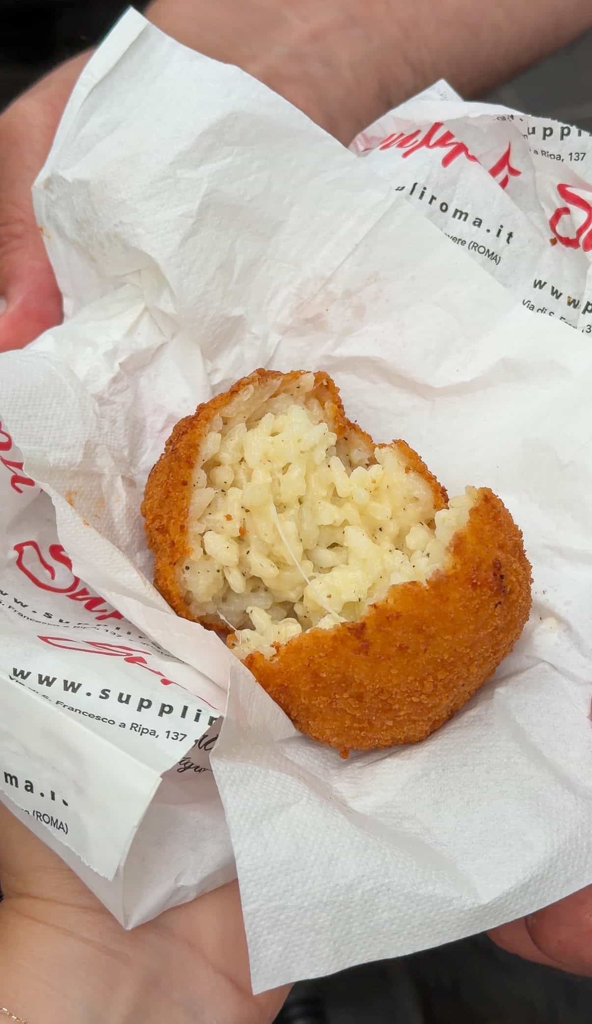 A hand holds a half-eaten, golden-brown fried rice ball with a creamy rice filling, wrapped in a white napkin. The rice ball is resting on a branded paper with red and black text.