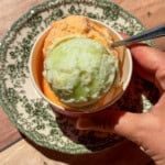 A hand holds a spoon in a bowl with two scoops of ice cream—one green and one orange, set on a decorative plate atop a wooden surface. This refreshing treat features Viral Melon Ice Cream (2 Ingredients), making it as simple as it is delightful.