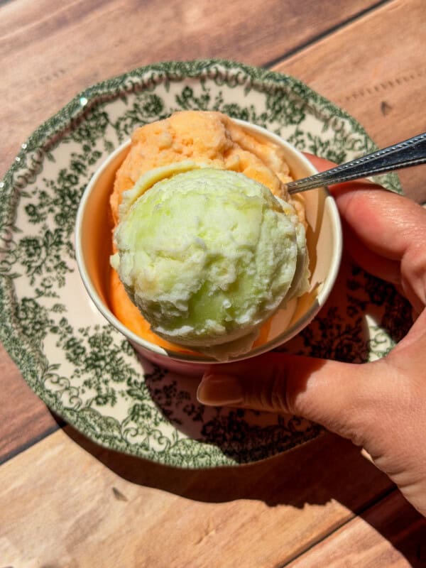 A hand holds a spoon in a bowl with two scoops of ice cream—one green and one orange, set on a decorative plate atop a wooden surface. This refreshing treat features Viral Melon Ice Cream (2 Ingredients), making it as simple as it is delightful.