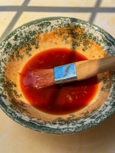 A paintbrush rests in a bowl of red sauce with shredded bits, placed on a table lit by sunlight. The ornate bowl, reminiscent of those used for serving Viral Melon Ice Cream (2 Ingredients), features a green floral pattern on the rim.