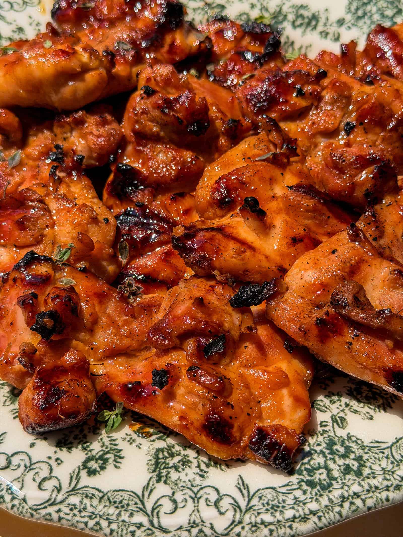 A close-up of grilled, charred chicken pieces with a reddish glaze served on a floral-patterned plate—perfect for pairing with a refreshing Viral Melon Ice Cream (2 Ingredients) dessert.